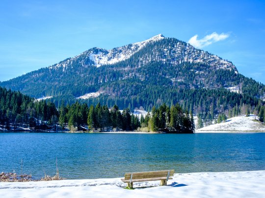 Mit einem Lächeln aufstehen im Hotel am Spitzingsee Bank am schneebedeckten See mit bewaldetem Berg im Hintergrund
