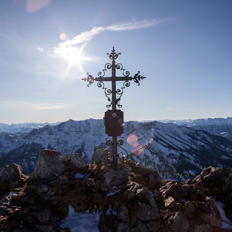 A look at Alte Wurzhütte Summit cross on rocky mountain with snowy Alps and sun in clear sky