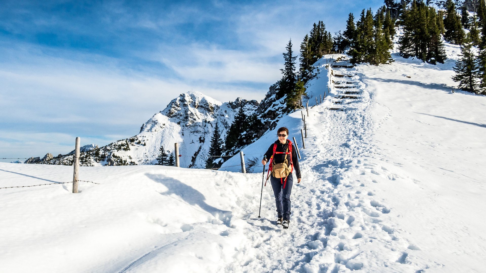 Woman hiking on snowy mountain trail with mountains and trees in the background