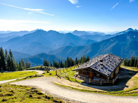 Mit einem Lächeln aufstehen im Hotel am Spitzingsee Almhütte mit Bergpanorama und geschwungenem Wanderweg bei klarem Himmel