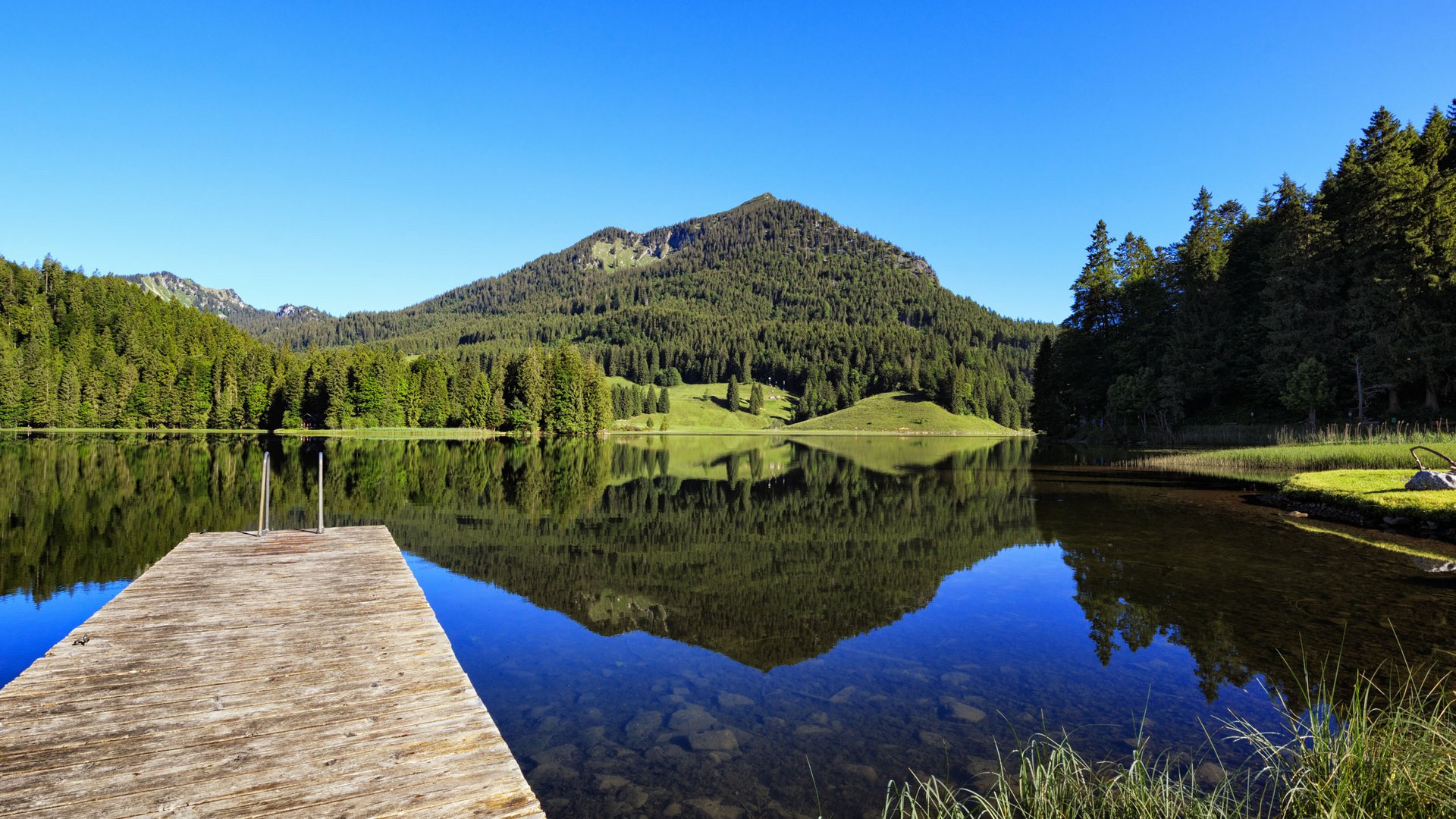 Steg an einem ruhigen Bergsee mit Wälder- und Bergreflexion bei klarem Himmel