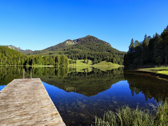 Mit einem Lächeln aufstehen im Hotel am Spitzingsee Steg an einem ruhigen Bergsee mit Wälder- und Bergreflexion bei klarem Himmel