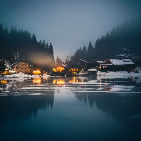 A look at Alte Wurzhütte Village by frozen lake at night with lit houses and snowy forest